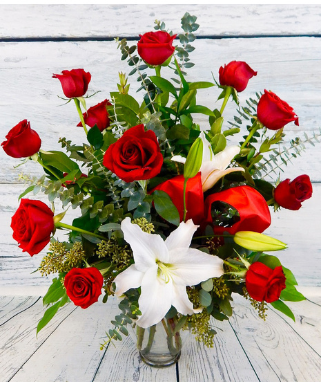 One dozen red roses with lilies and fancy greenery in an elegant, tall vase